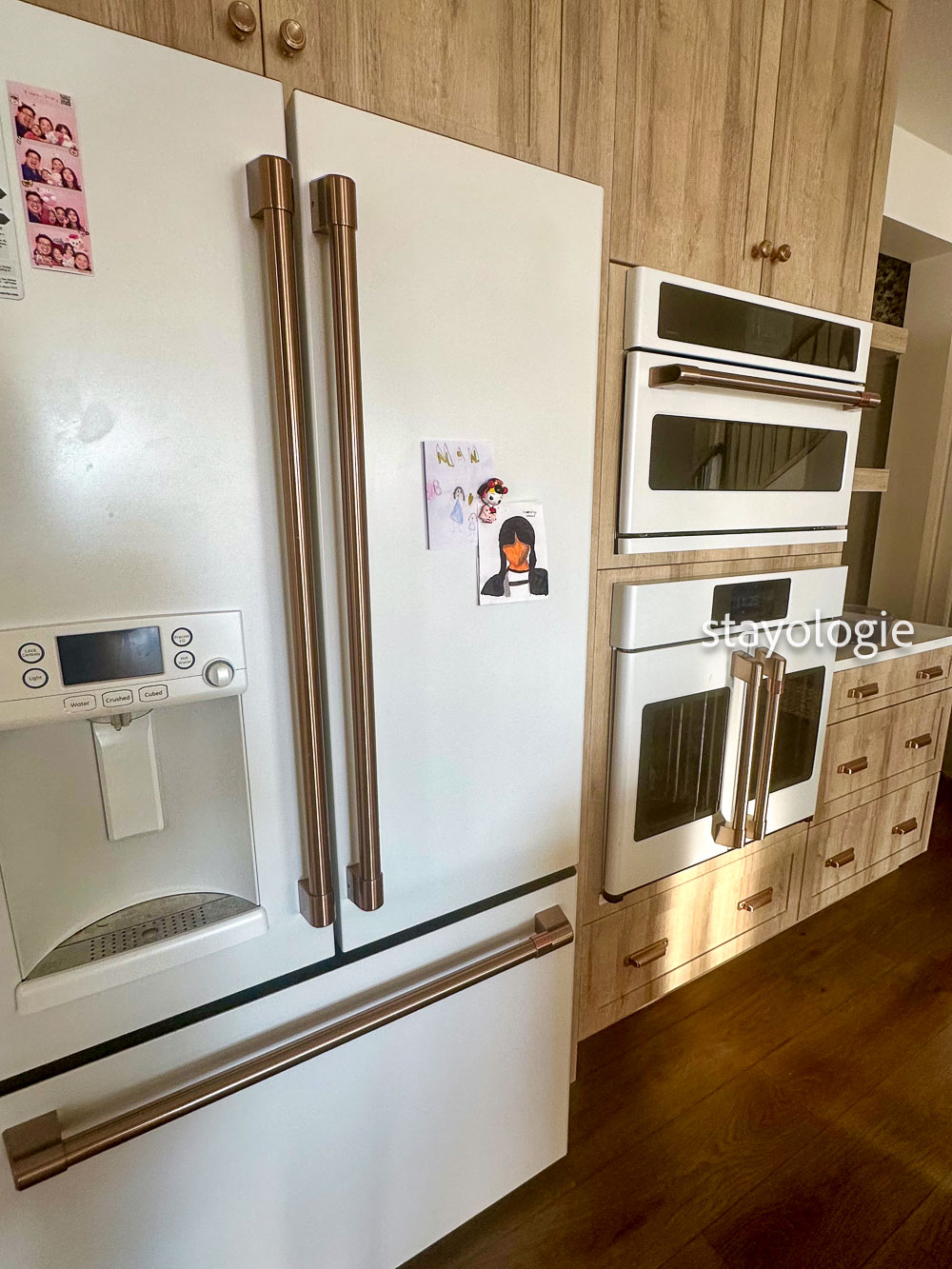 Kitchen with a white refrigerator displaying children’s artwork and family photos, surrounded by warm wood cabinetry and built-in appliances in a lived-in family home.